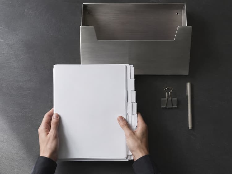 Overhead view of hands sliding a neat document stack toward a wall-mounted metal inbox with blank labels, suggesting the correct corporate submission channel