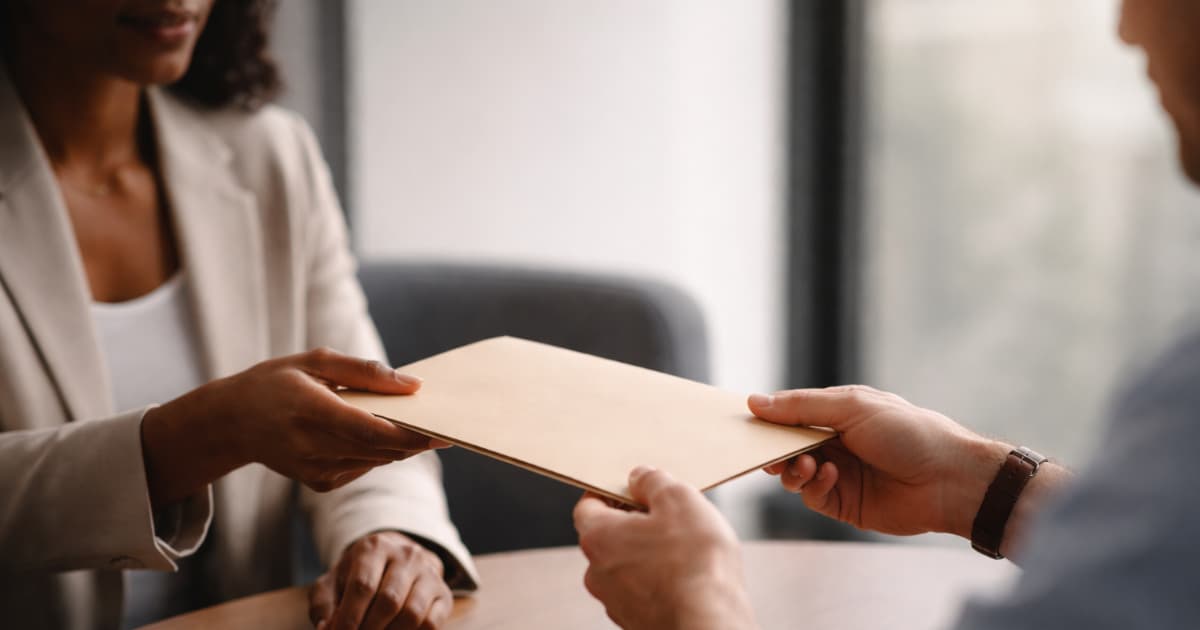 Two professionals exchanging a slim document folder across a small table in a modern office, illustrating client-freelancer tax paperwork handoff