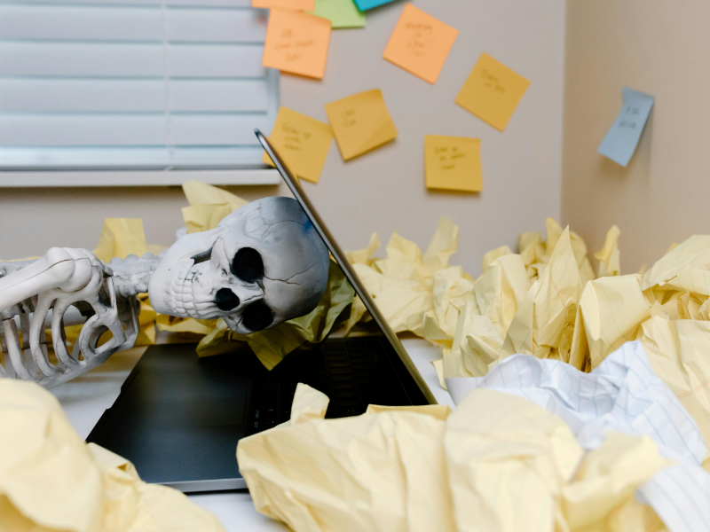 Skeleton at desk buried in crumpled papers and sticky notes still waiting for invoice payment