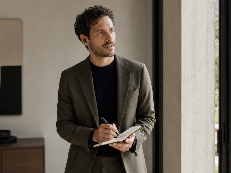 A freelancer on a headset jotting concise notes near a bright window, communicating calmly with a client about an invoice correction